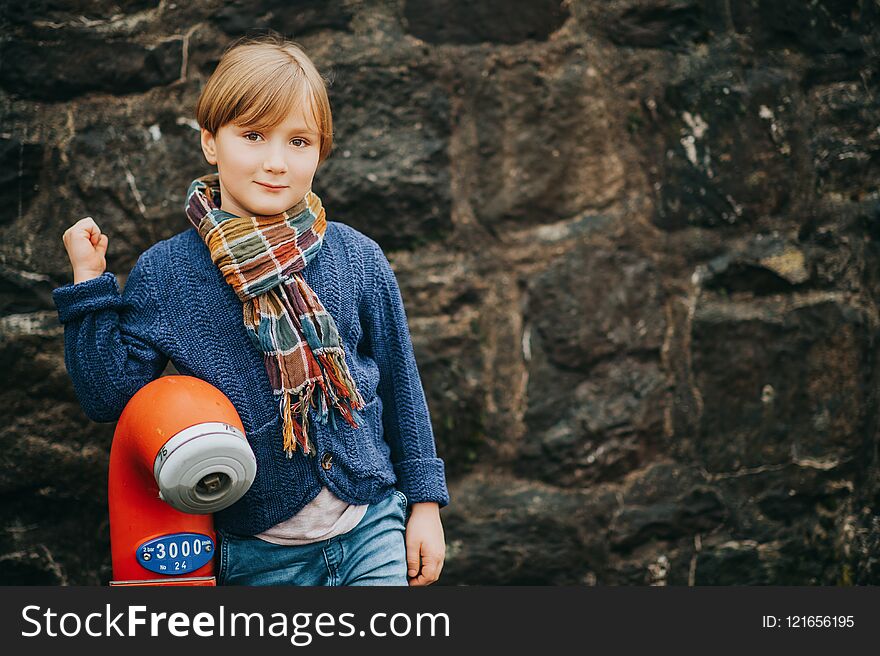 Outdoor portrait of handsome little boy wearing denim jeans, blue knitted jacket, stylish kid posing against dark wall. Fashion for small children