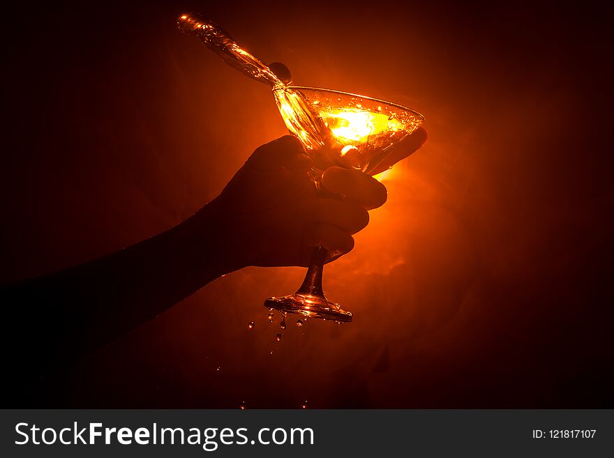 Martini cocktail glass in hand splashing on dark toned smoky background or colorful cocktail in glass with splashes and olives. Pa