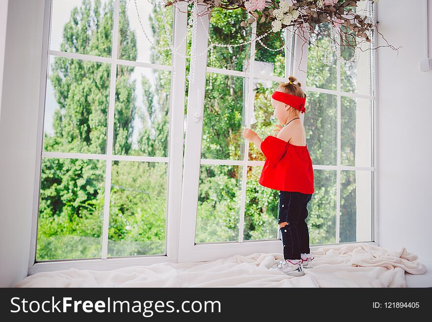 Little funny two years old baby girl in stylish red clothes and jeans and sneakers stands with back on the windowsill near the window. Headband and hairstyle on two heads in summer in sunny weather.