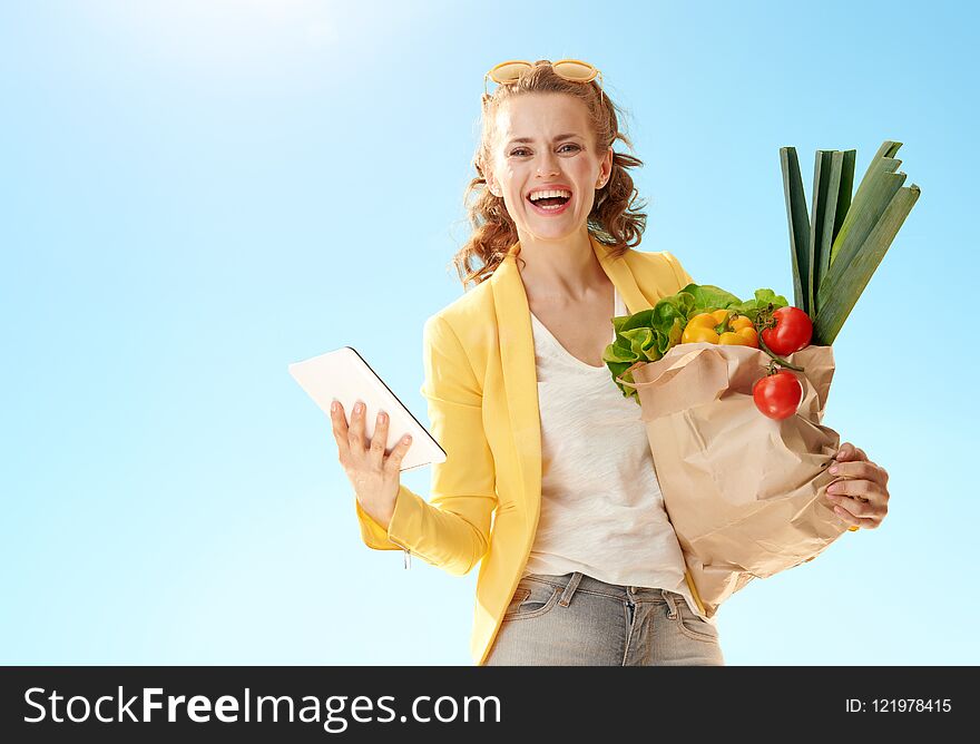 Smiling stylish woman in yellow jacket with paper bag with groceries with tablet PC against blue sky. Smiling stylish woman in yellow jacket with paper bag with groceries with tablet PC against blue sky