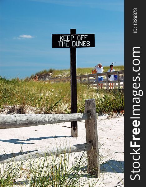 Beach scene with fence and sign that says Keep off the Dunes. There is sand, grass and a blue sky. There are people visible in the blurred background. Beach scene with fence and sign that says Keep off the Dunes. There is sand, grass and a blue sky. There are people visible in the blurred background.