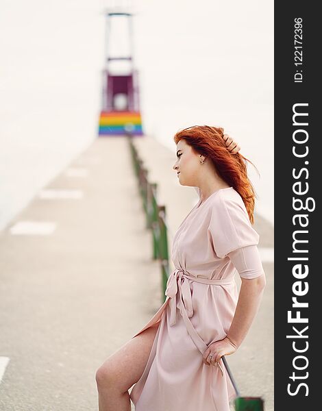 A young redheaded girl in a large round hat and pink dress sitting on the moorage near ocean. A young redheaded girl in a large round hat and pink dress sitting on the moorage near ocean