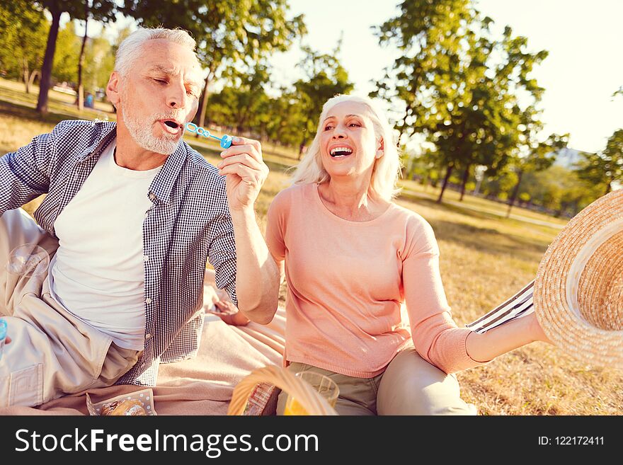 Never bored together. Emotional elderly women grinning broadly while looking at her husband blowing soap bubbles during a family picnic. Never bored together. Emotional elderly women grinning broadly while looking at her husband blowing soap bubbles during a family picnic.