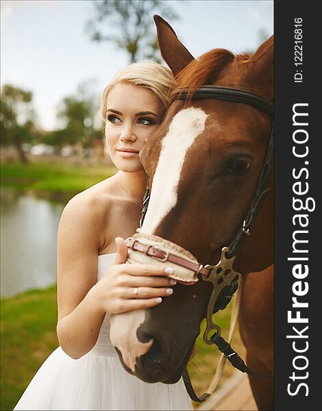 Beautiful and fashionable blonde model girl with blue eyes, with stylish hairstyle and bright makeup, in white dress posing with brown horse outdoors.