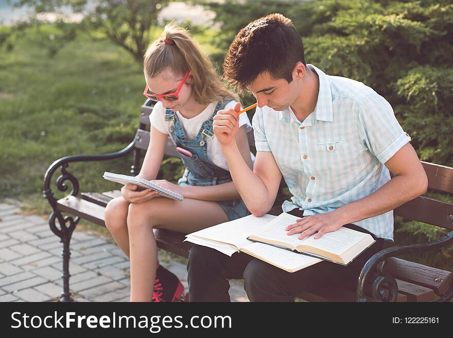 Students making the notes learning from books sitting on a bench in a park. Young boy wearing a blue shirt and dark jeans. Young blondie girl wearing jeans and sunglasses. Students making the notes learning from books sitting on a bench in a park. Young boy wearing a blue shirt and dark jeans. Young blondie girl wearing jeans and sunglasses