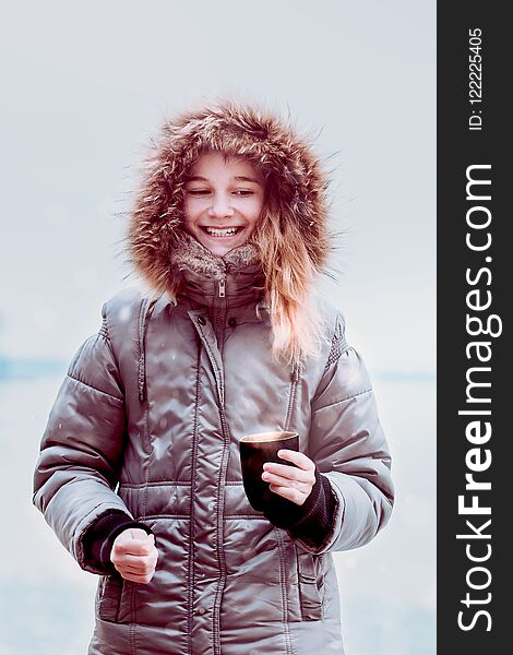 Young pretty smiling girl enjoying drinking hot drink during walk on a wintery day. Wearing coat with hood. Hold a mug of tea. Young pretty smiling girl enjoying drinking hot drink during walk on a wintery day. Wearing coat with hood. Hold a mug of tea