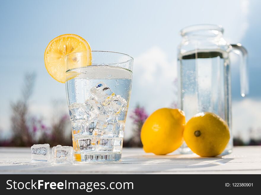 Glass of ice and water decorated with a slice of lemon standing on a white table against a decanter with water and two lemons. Beautiful still life of soft drinks.