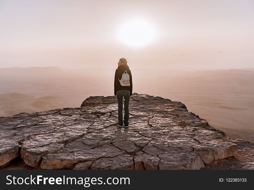 Alone young girl with backpack in israel negev desert admires the view of sunrise. Young female person stands on the edge of the cliff of makhtesh ramon park. Beautiful horizon landscape