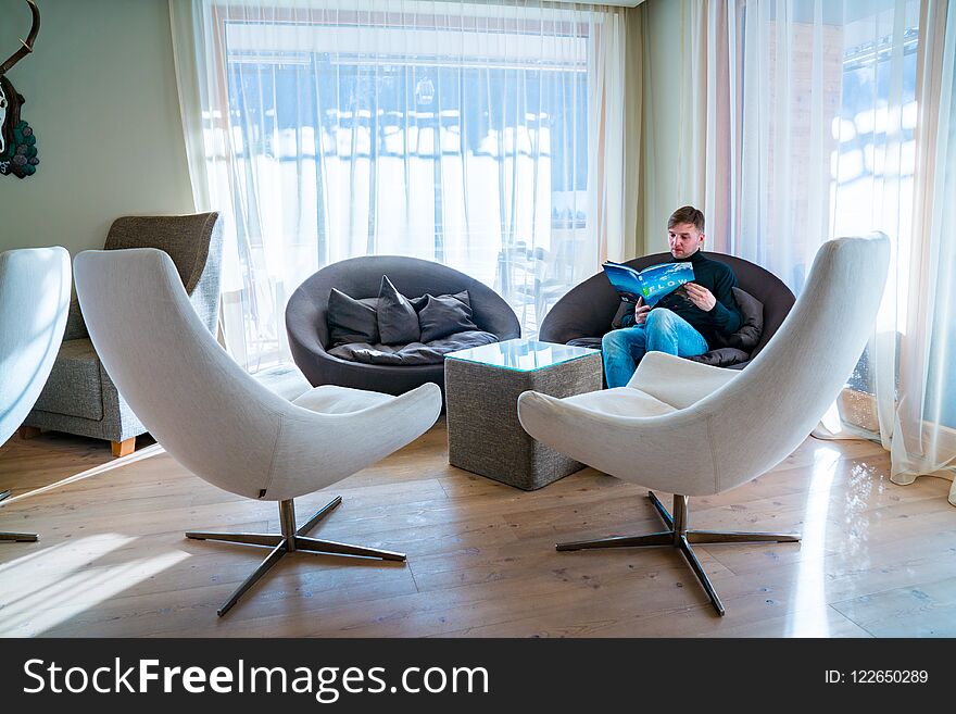 Saalbach, Austria. Alpina hotel. March 20, 2018. Young man sitting in a chair reading a book in a comfortable library room near huge windows.