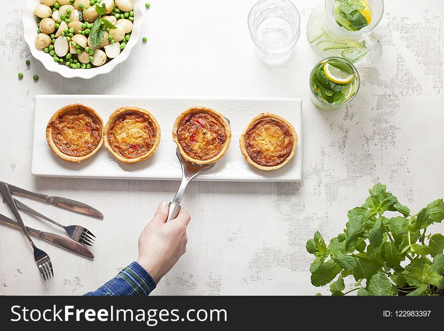 A Chef places a mini quiche on a white plate with three others on a white table with cutlery and a dish of peas and potatoes. A Chef places a mini quiche on a white plate with three others on a white table with cutlery and a dish of peas and potatoes