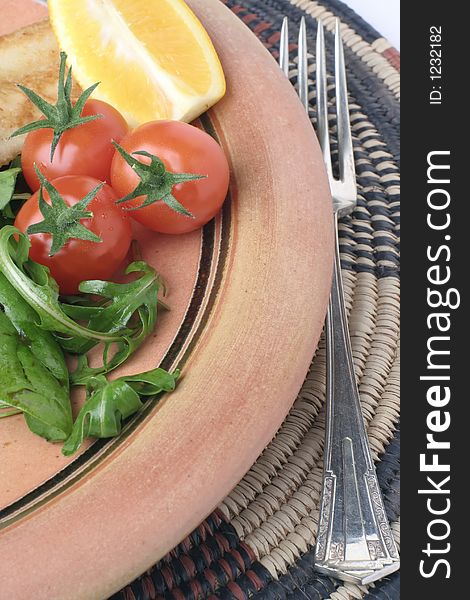 A glazed stoneware plate with Salad and fork on an African woven table mat. Some grilled fish in the background. A glazed stoneware plate with Salad and fork on an African woven table mat. Some grilled fish in the background.