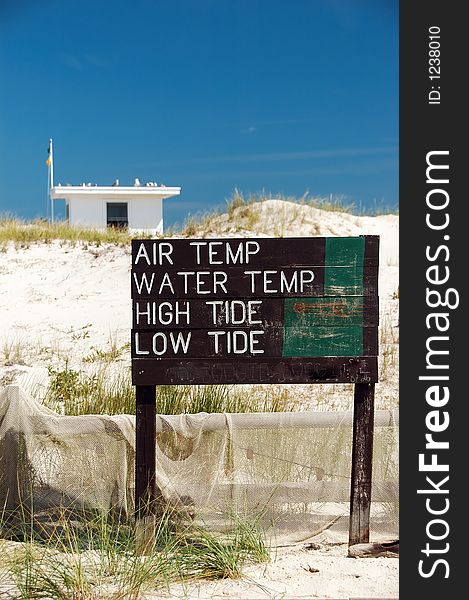 A beach scene with a fence, net and water conditions sign.  A lifeguard shack, flags and seagulls are in blurred background.  The sky is blue almost cloudless. A beach scene with a fence, net and water conditions sign.  A lifeguard shack, flags and seagulls are in blurred background.  The sky is blue almost cloudless.