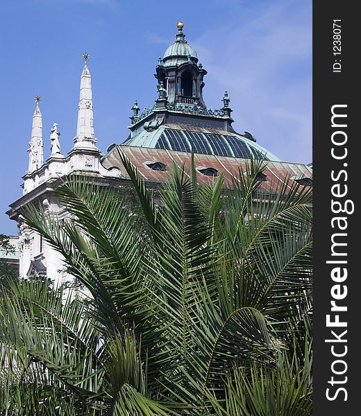 The image shows an old palace in the center of Munich. In front of the building is a palm tree. In the background of the picture is blue sky. The image shows an old palace in the center of Munich. In front of the building is a palm tree. In the background of the picture is blue sky.