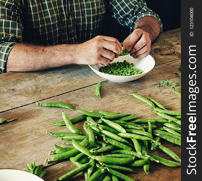 Male hands clean green peas sitting at wooden table in the kitchen. Rustic style. Green peas in bowl on wooden table. Male hands clean green peas sitting at wooden table in the kitchen. Rustic style. Green peas in bowl on wooden table