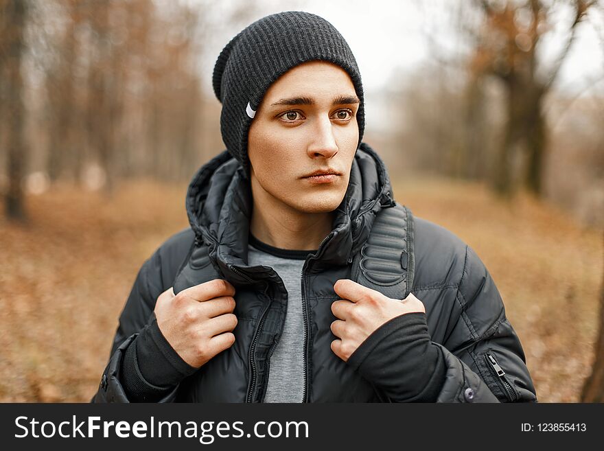 Stylish young man in a black knitted hat and jacket standing near the autumn trees.