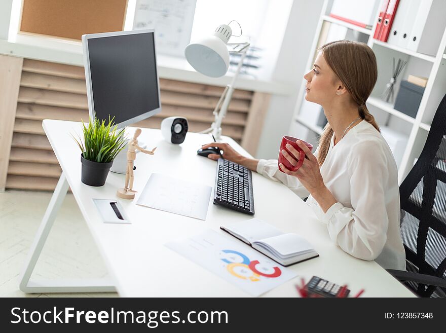 A young girl is sitting at a table in the office, holding a red cup in her hand and working at the computer.