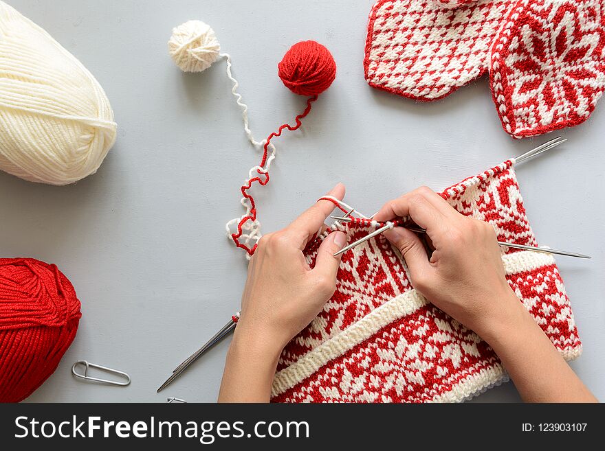 Girl knits red and white Norwegian jacquard hat knitting needles on gray wooden background. Process of knitting. Top view. Flat lay