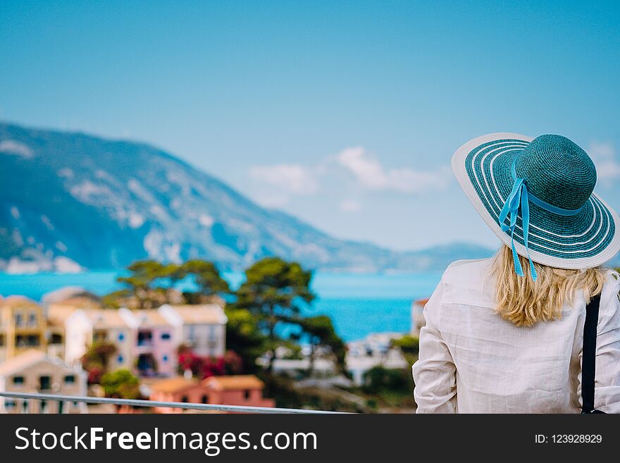 Tourist woman wear blue sunhat and white clothes admire view of colorful tranquil village Assos on sunny day. Stylish female visiting Kefalonia enjoying summer time on Greece travel vacation.