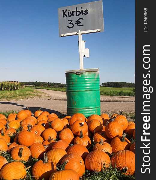 The image shows a pile of orange pumpkins (in German it is 'KÃƒÂ¼rbis') before a green barrel. On top of the barrel is a silver sign, showing that a pumpkin costs 3 Euro. Behind the barrel is a small road. The sky in the picture is blue. The cucurbits are mainly sold as halloween decoration. The image shows a pile of orange pumpkins (in German it is 'KÃƒÂ¼rbis') before a green barrel. On top of the barrel is a silver sign, showing that a pumpkin costs 3 Euro. Behind the barrel is a small road. The sky in the picture is blue. The cucurbits are mainly sold as halloween decoration.