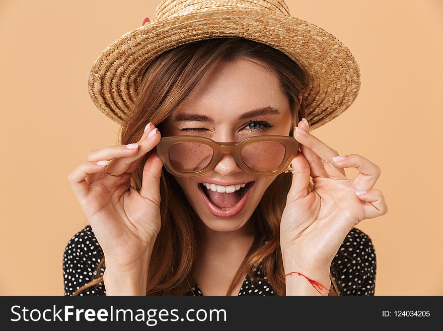 Image of summer pretty woman 20s wearing straw hat touching sunglasses and smiling at camera over beige background