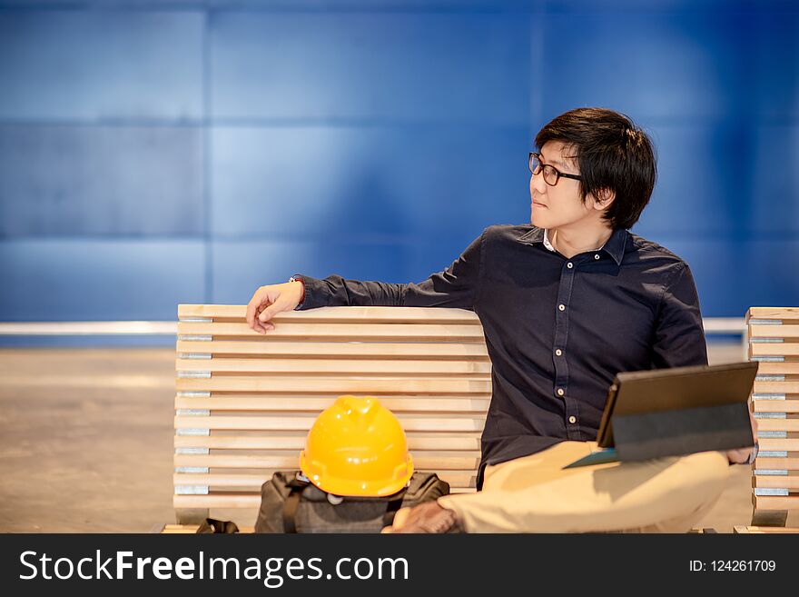 Young Asian engineer man using digital tablet while sitting with personal yellow protective safety helmet and bag on wooden bench. Male architect or Engineering concepts. Young Asian engineer man using digital tablet while sitting with personal yellow protective safety helmet and bag on wooden bench. Male architect or Engineering concepts