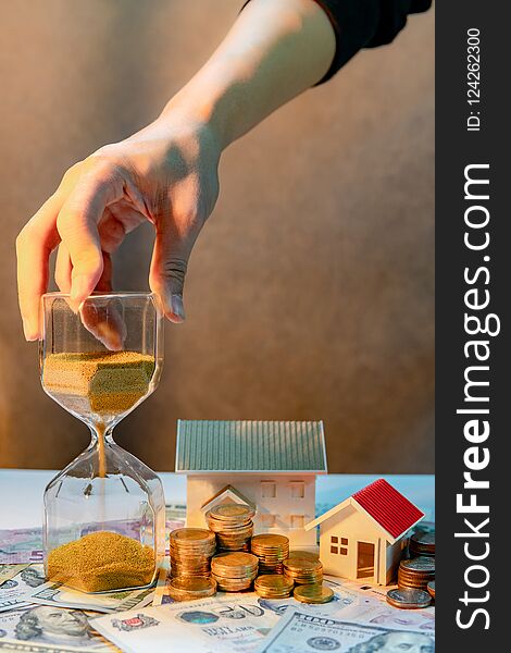 Real estate or property investment growing business. Home mortgage loan rate. Saving money for retirement concept. Male hand holding hourglass with house model and coin stack on banknotes on the table. Real estate or property investment growing business. Home mortgage loan rate. Saving money for retirement concept. Male hand holding hourglass with house model and coin stack on banknotes on the table