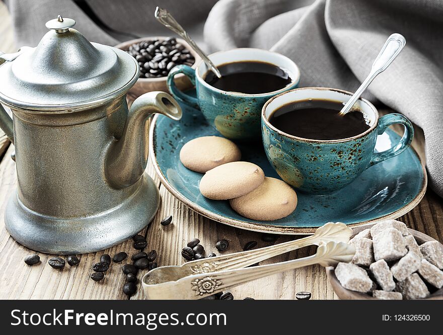 Two blue vintage cups of black coffee, biscuits and antique pewter coffee pot surrounded by linen cloth, sugar pieces and coffee beans on old wooden table. Two blue vintage cups of black coffee, biscuits and antique pewter coffee pot surrounded by linen cloth, sugar pieces and coffee beans on old wooden table