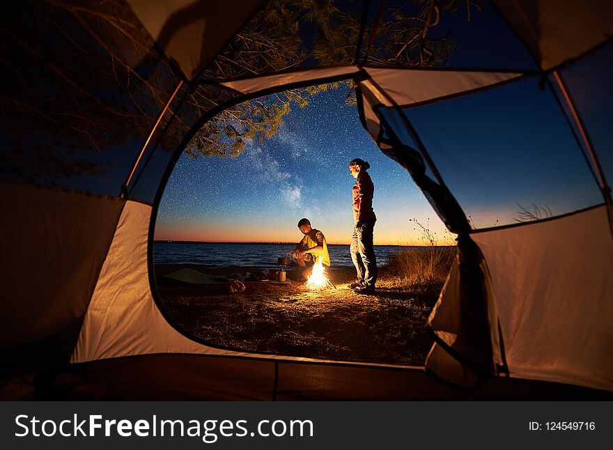 Camping on lake shore at sunset, view from inside tourist tent. Young romantic couple, man and woman preparing dinner on campfire on blue sea water background. Tourism, recreation and love concept. Camping on lake shore at sunset, view from inside tourist tent. Young romantic couple, man and woman preparing dinner on campfire on blue sea water background. Tourism, recreation and love concept.