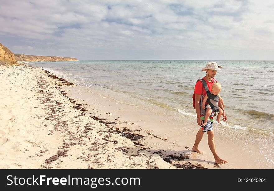 Father and his infant son in baby carrier walk on the sea beach.
