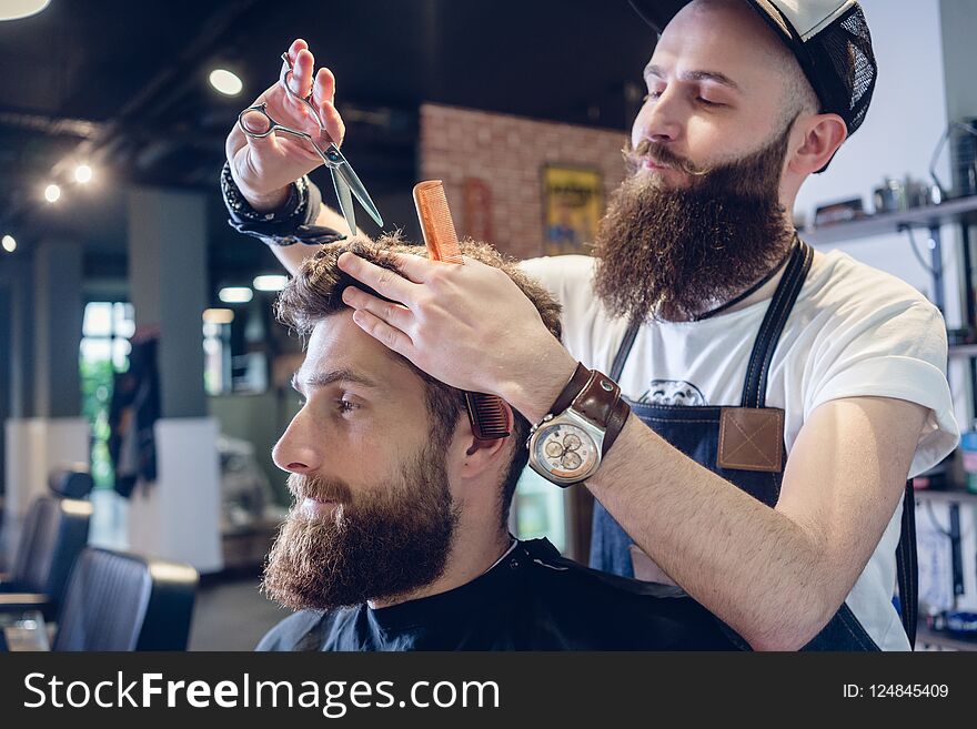 Dedicated male hairstylist using scissors and plastic comb while giving a cool haircut to a redhead bearded young men in a trendy beauty salon. Dedicated male hairstylist using scissors and plastic comb while giving a cool haircut to a redhead bearded young men in a trendy beauty salon