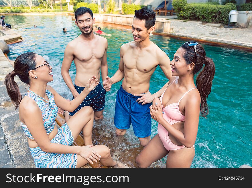 Multi-ethnic group of four young and cheerful friends smiling and talking while relaxing together at a trendy outdoor swimming pool in summer. Multi-ethnic group of four young and cheerful friends smiling and talking while relaxing together at a trendy outdoor swimming pool in summer