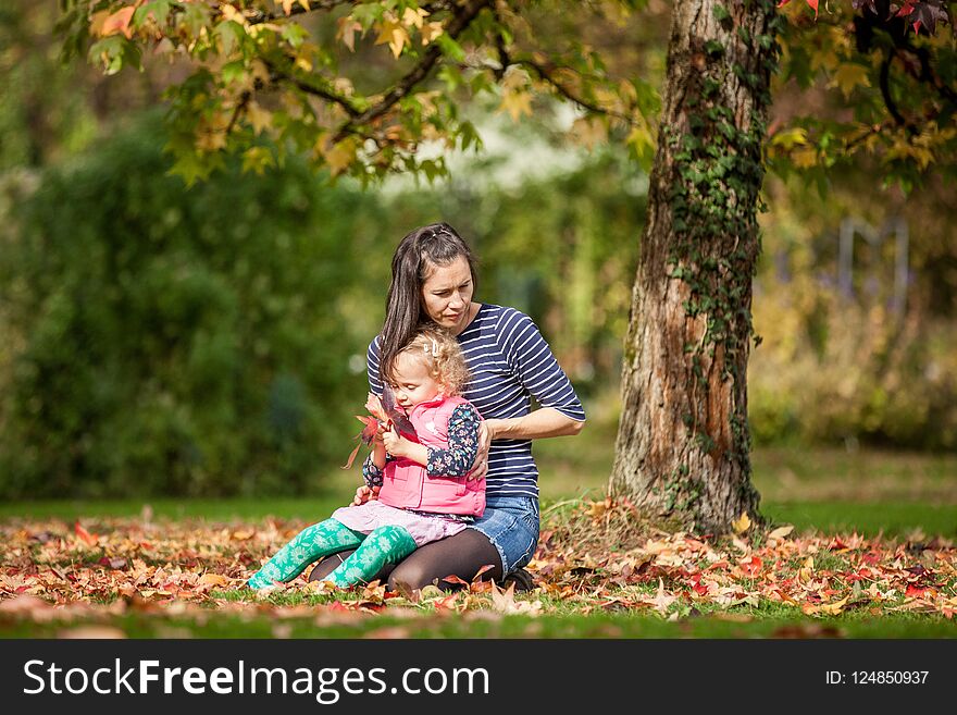 Mother and girl having fun under tree with autumn leaves in the park, blond cute curly girl, happy and young family