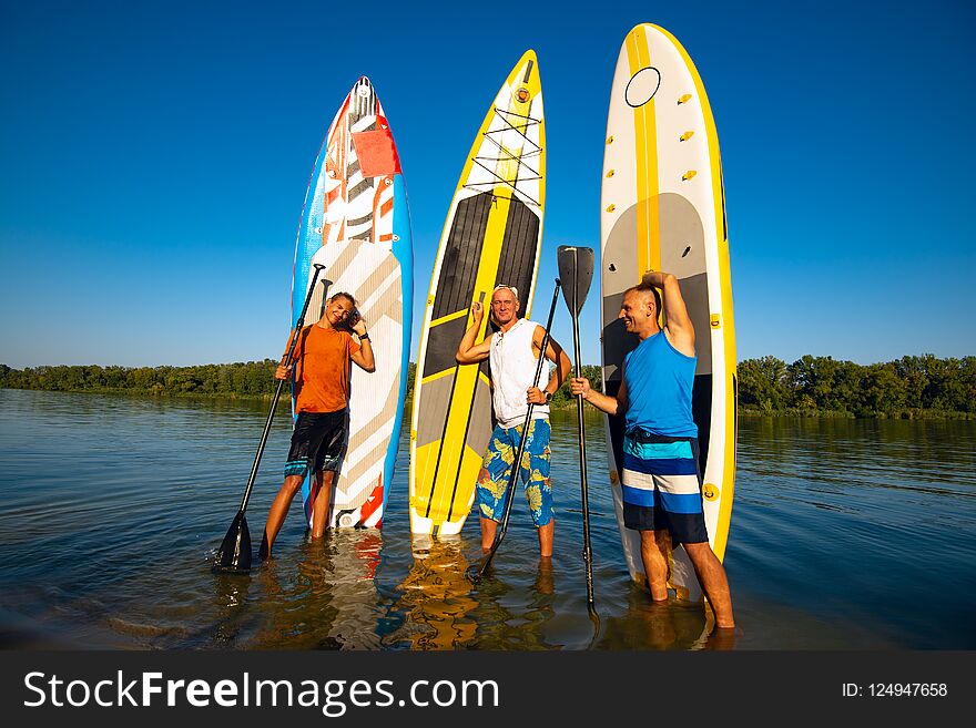 Joyful friends with SUP boards in their hands stand on the bank of a big river in the summer evening and enjoy life.