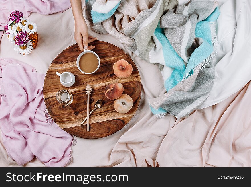 Cozy autumn or winter flatlay of wooden tray with cup of coffee, peaches, creamer with plant milk, syrup and spoons on pastel sheets and woman`s hand, selective focus