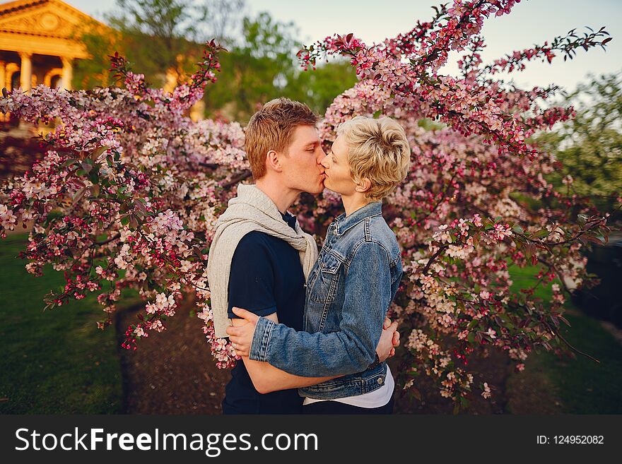 Stylish and beautiful women with short light hair, dressed in a blue jeans jacket walks with her handsome men in a sunny park and standind near flower trees. Stylish and beautiful women with short light hair, dressed in a blue jeans jacket walks with her handsome men in a sunny park and standind near flower trees