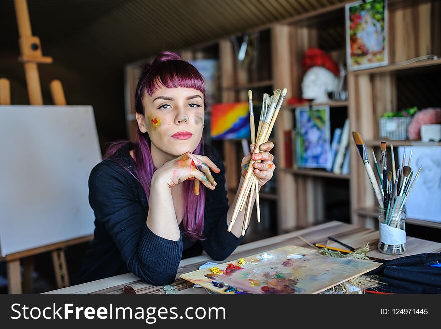 Beautiful and serious female artist with purple hair and dirty hands with different paints on them, holding paintbrushes in her art studio.