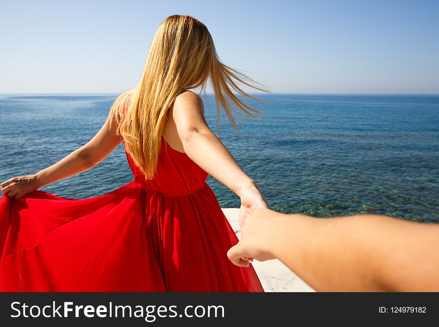 Follow me. Young blond woman in the red dress holding the friend`s hand at the beach in Cyprus.