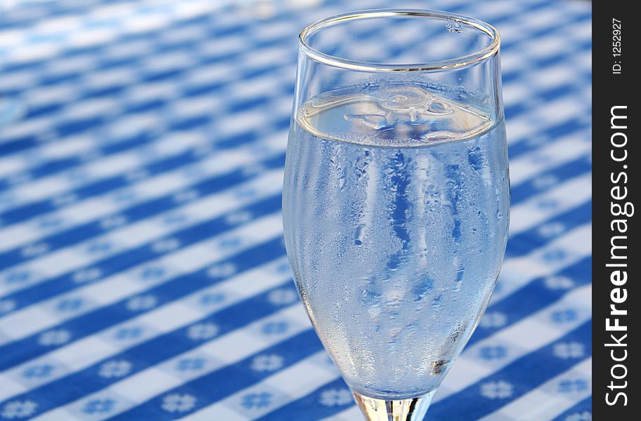A glass of iced water, standing on a blue and white tablecloth, ice cubes in glass and condensed water on glass due to heat. A glass of iced water, standing on a blue and white tablecloth, ice cubes in glass and condensed water on glass due to heat