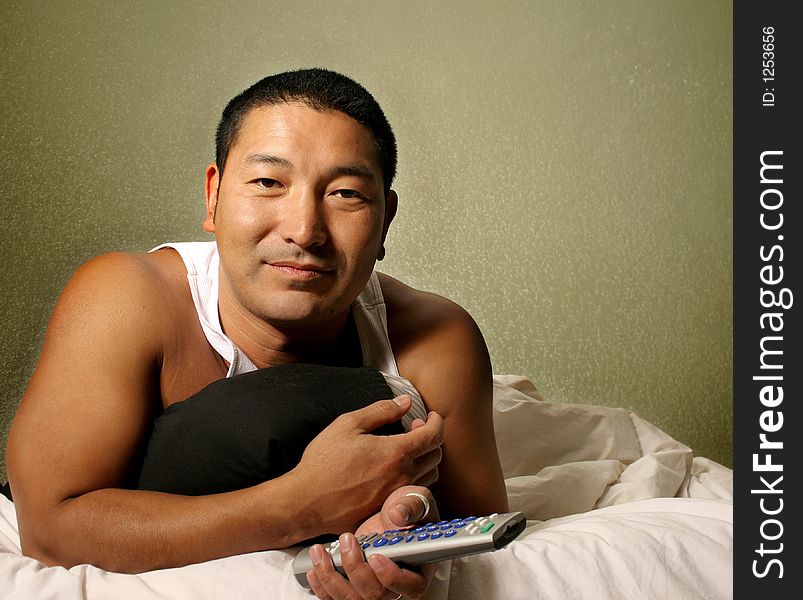 Asian man laying in bed holding a remote control. Asian man laying in bed holding a remote control
