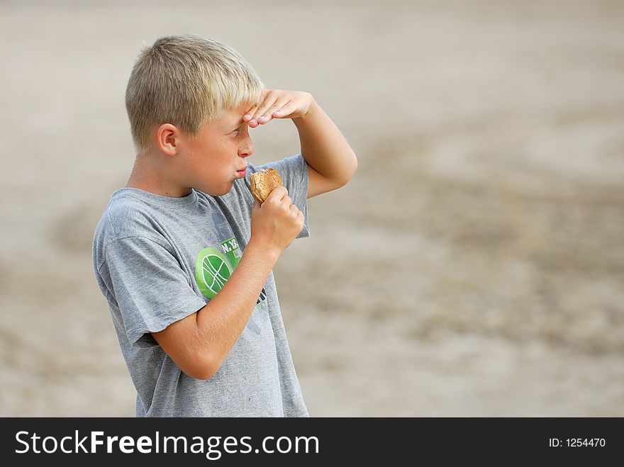 Young boy is watching the scenery at the beach and is eating a hamburger. Young boy is watching the scenery at the beach and is eating a hamburger