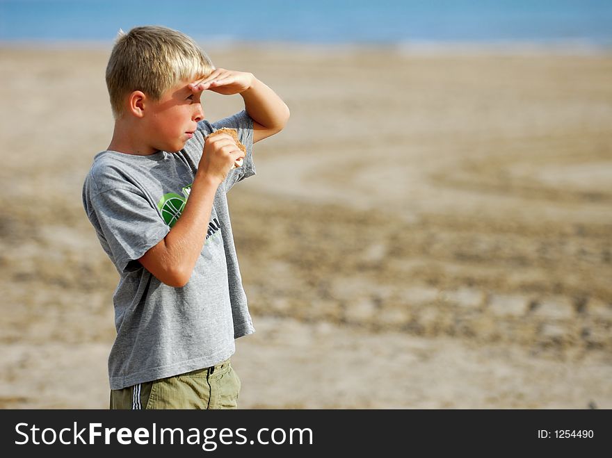 Young boy is watching the scenery at the beach and is eating a hamburger. Young boy is watching the scenery at the beach and is eating a hamburger