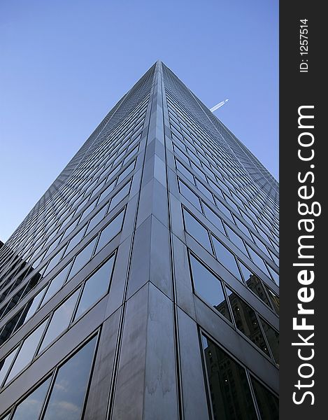 Upward view along the corner edge of a skyscraper against a bright blue sky background. A passing airplane is visible above the building. Upward view along the corner edge of a skyscraper against a bright blue sky background. A passing airplane is visible above the building.