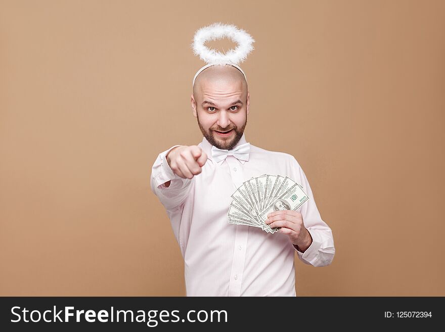 This is for you. Happy handsome middle aged bald bearded angel in shirt and white halo on head holding many dollars, pointing and looking at camera. studio shot, isolated on light brown background.