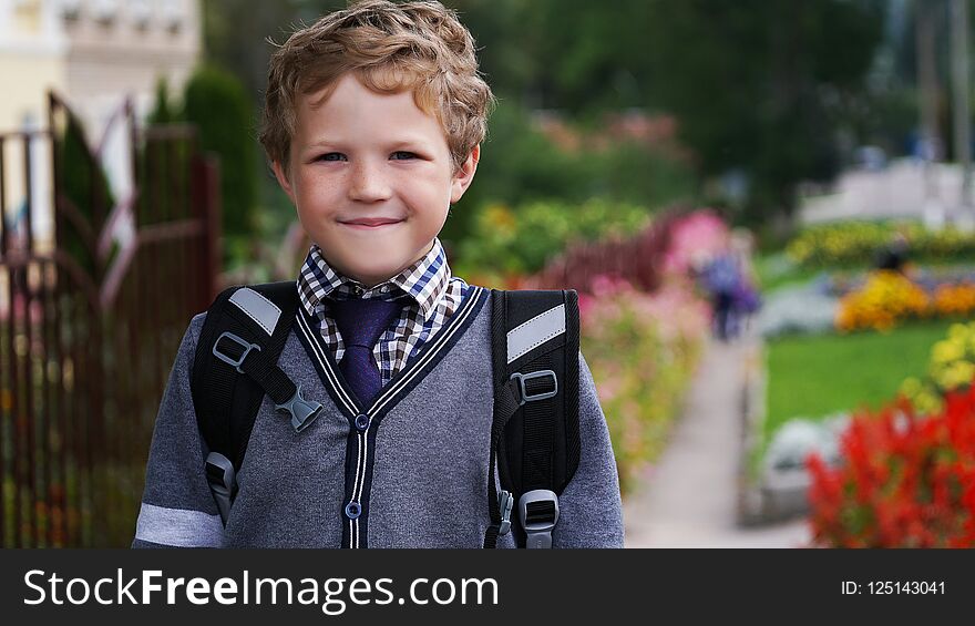 Smiling little curly kid boy with backpack or satchel on his first day to school or nursery. Child outdoors on warm sunny day, Back to school concept. School yard in color flowers. Smiling little curly kid boy with backpack or satchel on his first day to school or nursery. Child outdoors on warm sunny day, Back to school concept. School yard in color flowers.