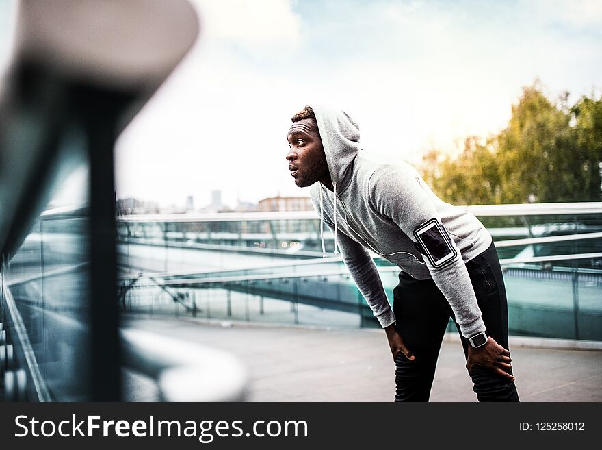 Young sporty black man runner with smartphone on the bridge in a city, resting.