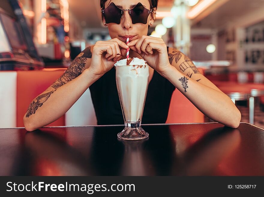 Woman wearing star shaped eyeglasses drinking milkshake at a restaurant. Woman with tattooed arms drinking milkshake with a straw at a diner. Woman wearing star shaped eyeglasses drinking milkshake at a restaurant. Woman with tattooed arms drinking milkshake with a straw at a diner.