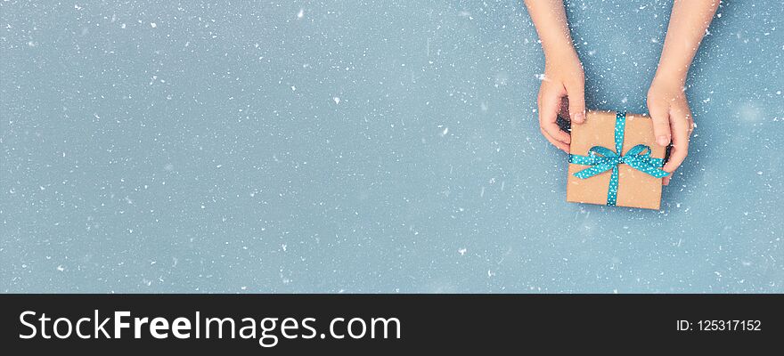 Child hands holding one gift box wrapped in kraft paper tied with blue ribbon in polka dots on blue grey background. Top view, place for text. Holiday concept, banner for site. Child hands holding one gift box wrapped in kraft paper tied with blue ribbon in polka dots on blue grey background. Top view, place for text. Holiday concept, banner for site.