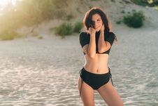 Young European Woman With Dark Thick Hair, Dressed In A Black Separate Swimsuit, Posing On White Sand At Sunset Stock Photos