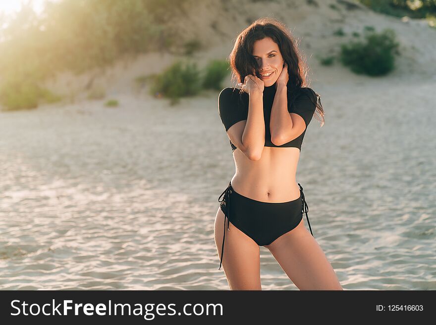 young European woman with dark thick hair, dressed in a black separate swimsuit, posing on white sand at sunset