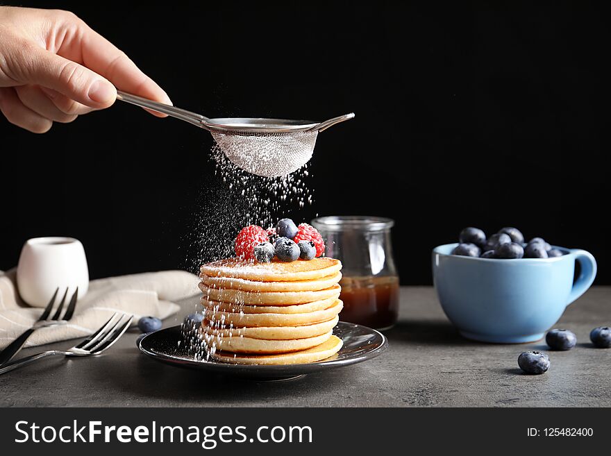 Woman adding sugar powder to tasty pancakes with berries on plate, closeup